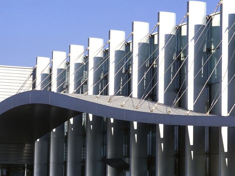 Facciata curva e moderna di un edificio con pilastri metallici e pannelli di vetro contro un cielo blu.
