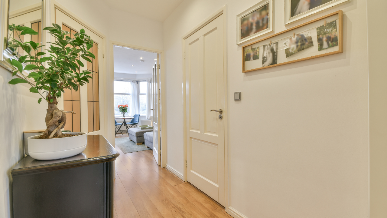 Hallway with wooden floor, a black cabinet with bonsai, and framed photos on white wall.