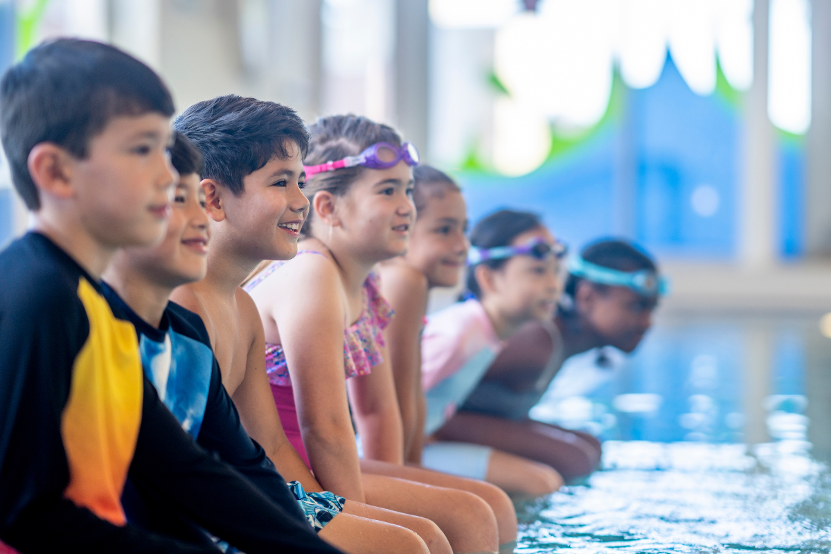 Children in swimsuits sit on the edge of a pool, looking at the water, smiling.
