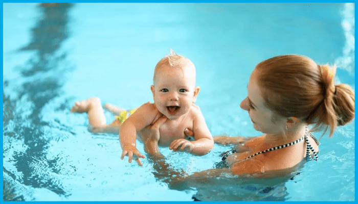 Mother and baby in a pool; the baby is smiling, and the mother is facing him, smiling.
