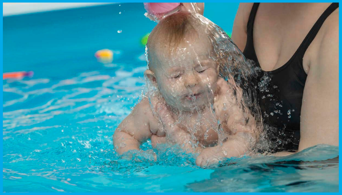 Baby in a swimming pool with eyes closed as water is gently poured over their face by a caregiver.