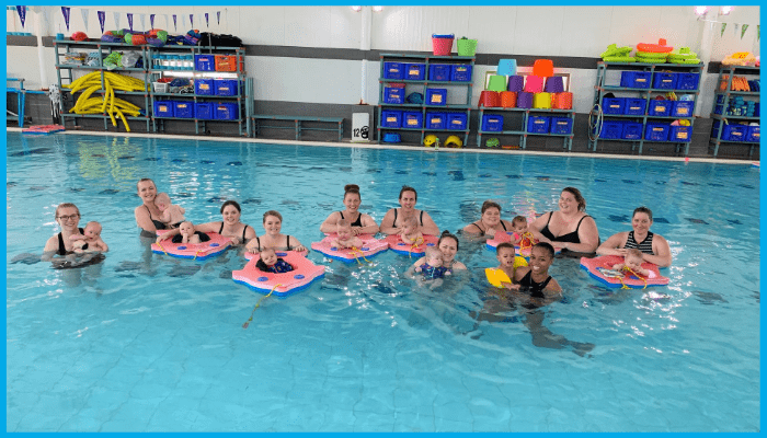 Parents and babies participating in a group swim class in a warm indoor pool at a Canberra swim school.
