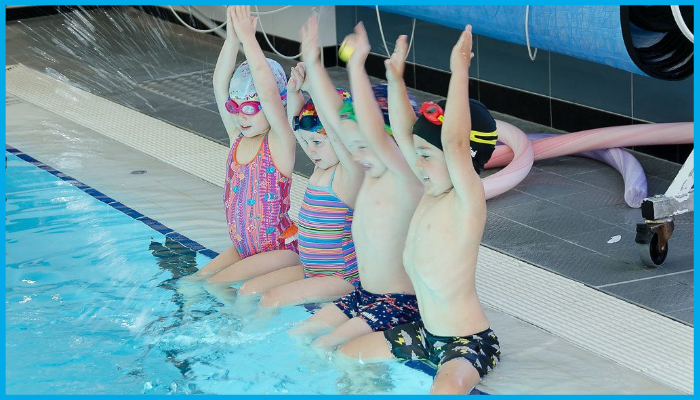 Children in colourful swimwear sitting poolside at a Canberra swimming lesson, raising arms to practise safe entry, supervised by certified instructors in a clean indoor pool facility.