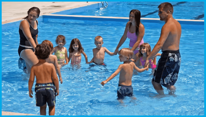 Group of children and adults standing in a circle in a shallow pool, preparing to play “Ring Around the Rosie.
