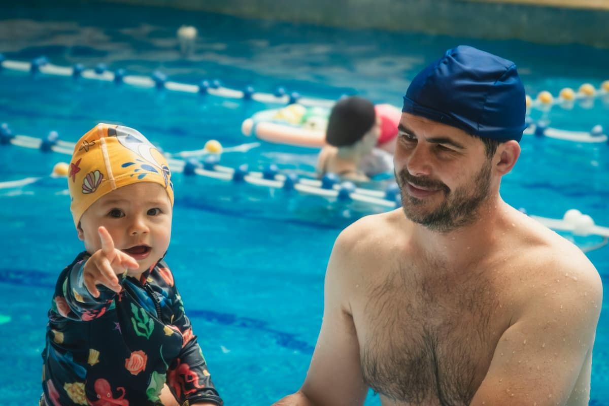 Father and child in a pool, smiling. Child points while wearing a swim suit and cap, father in swim cap.