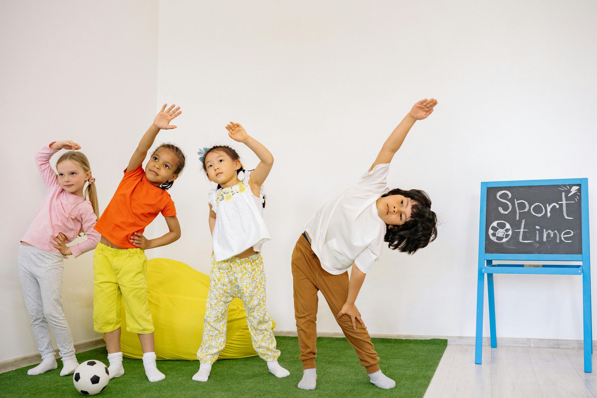 Four children stretching in a room. One child holds a soccer ball. A chalkboard displays 