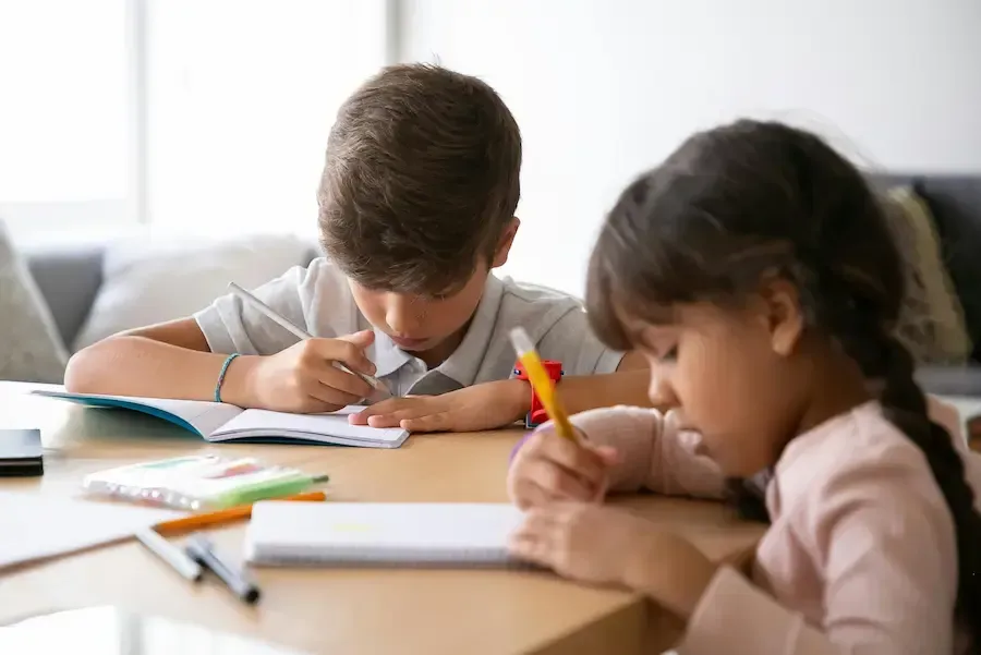 Two children writing in notebooks at a wooden table; pens and pencils are visible.