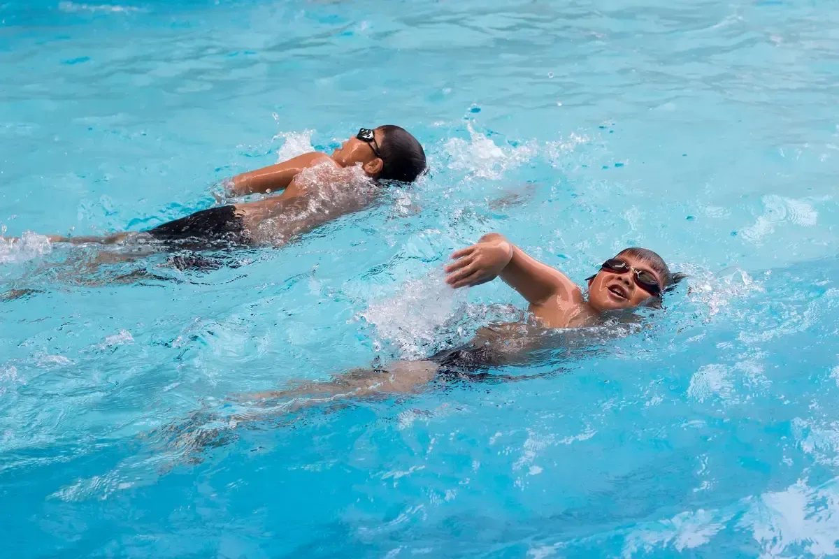 Two swimmers in a blue pool, one doing freestyle, the other backstroke. Water splashes.