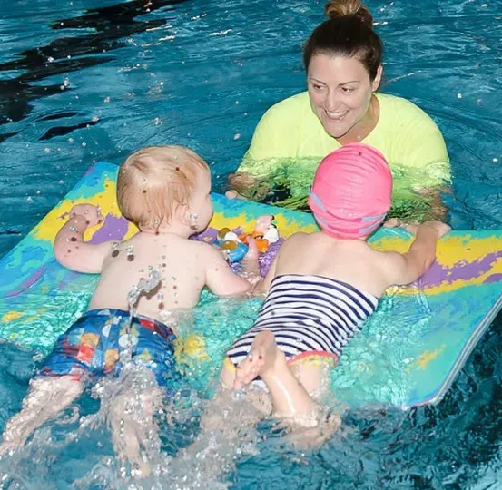 Alena Sarri and two toddlers on a floating mat in a swimming pool. The instructor smiles, watching them play.