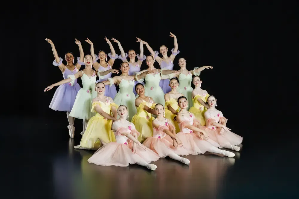 Ballet dancers in pastel tutus pose with arms raised against a black backdrop.