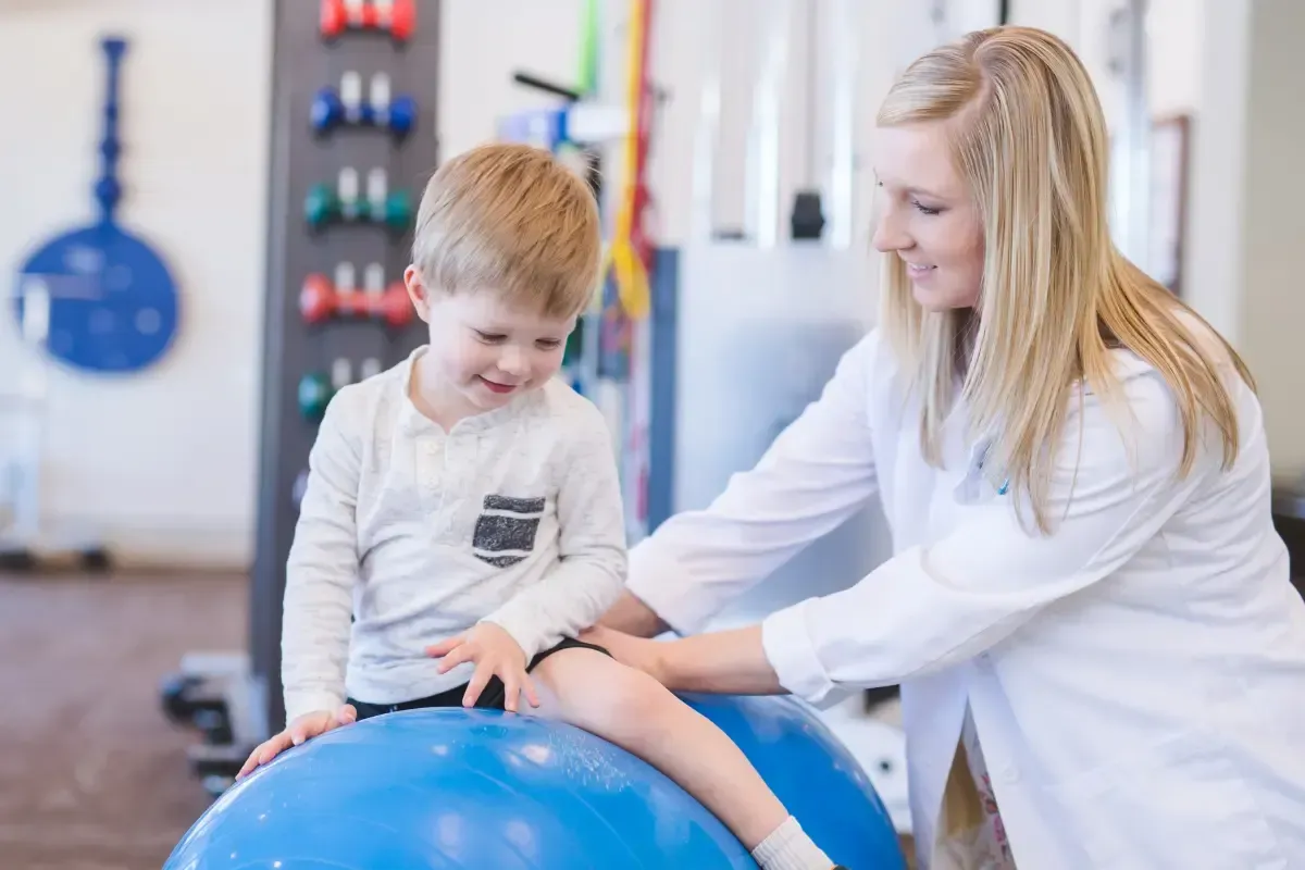 A child sits on a blue exercise ball, a healthcare provider gently holds his leg. Inside a clinic.