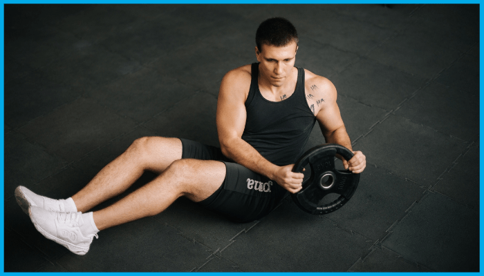 Man sitting on the floor, holding a weight plate, twisting his torso. He wears black athletic clothing and white sneakers.