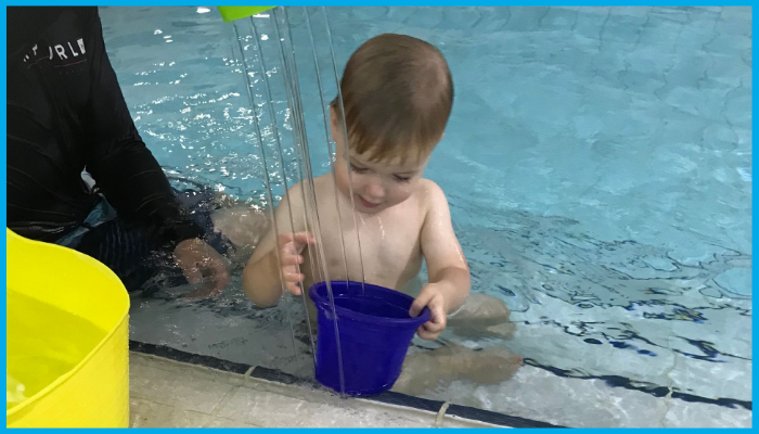 A child in a pool, playing with a blue bucket under water streams, with an adult nearby.