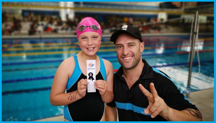 Swim coach celebrating with young competitive swimmer holding second place ribbon at poolside