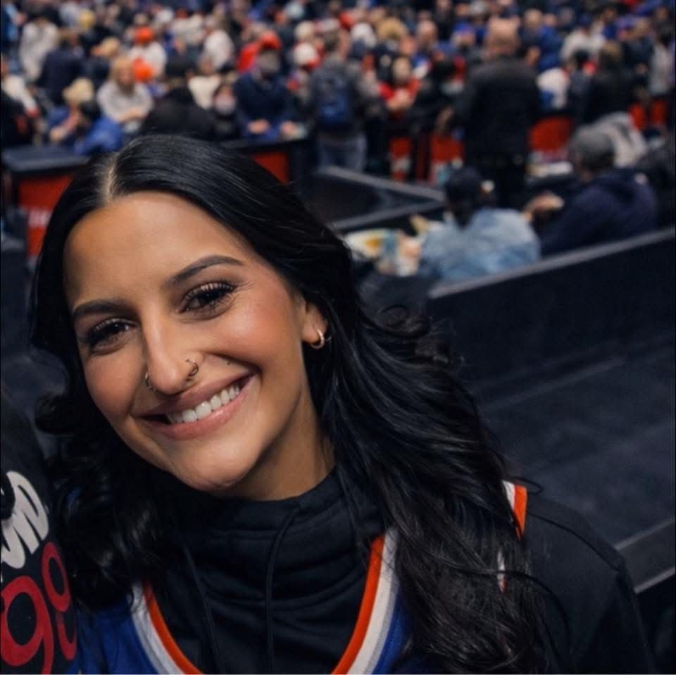 A smiling person with dark hair and a nose ring at a crowded sports arena, wearing a blue and orange jersey.