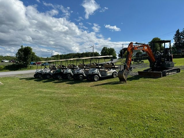A row of golf carts are parked in a grassy field next to an excavator.