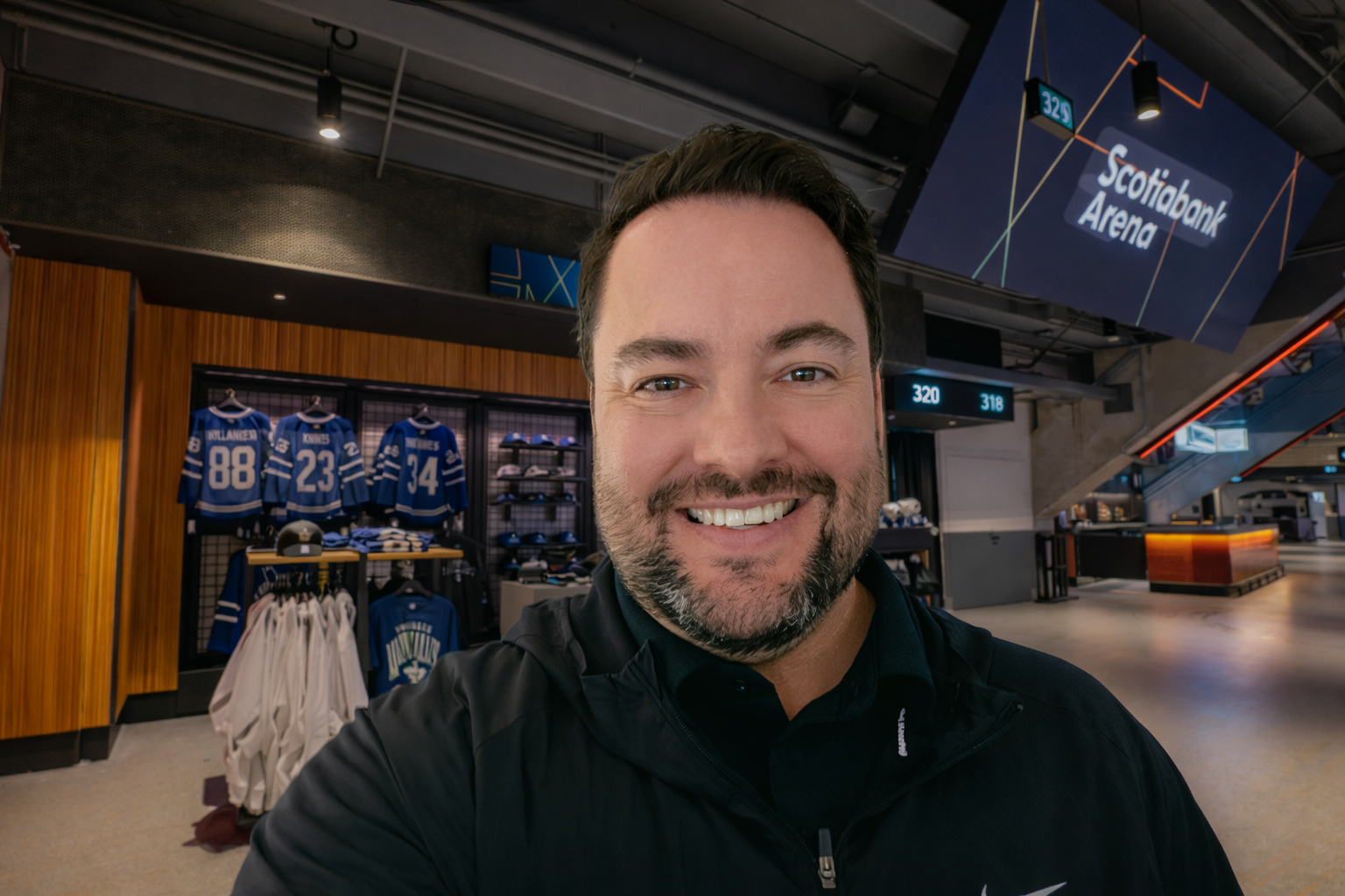 A man with a beard wearing a black shirt is smiling in front of a brick wall.