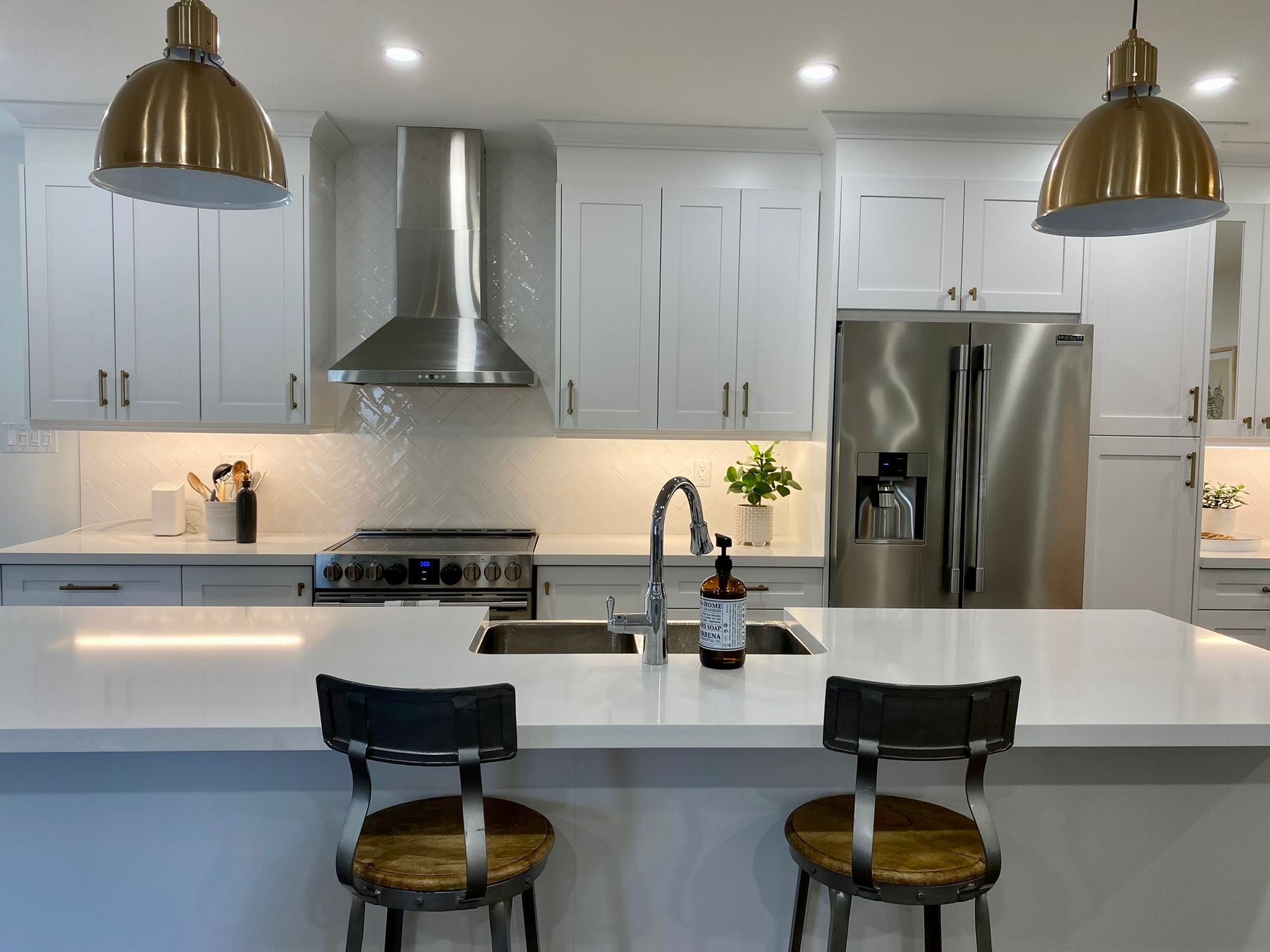 Modern white kitchen with stainless steel appliances, white countertops, and gold pendant lights. Two bar stools sit at an island.