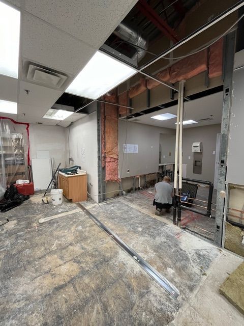 Room under construction with exposed walls and ceiling, a person working near pipes, debris on the floor.