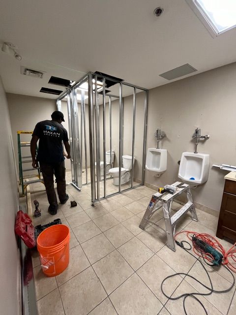 Bathroom renovation: Man near unfinished stalls, two urinals, and toilet. Tools and supplies on the floor.