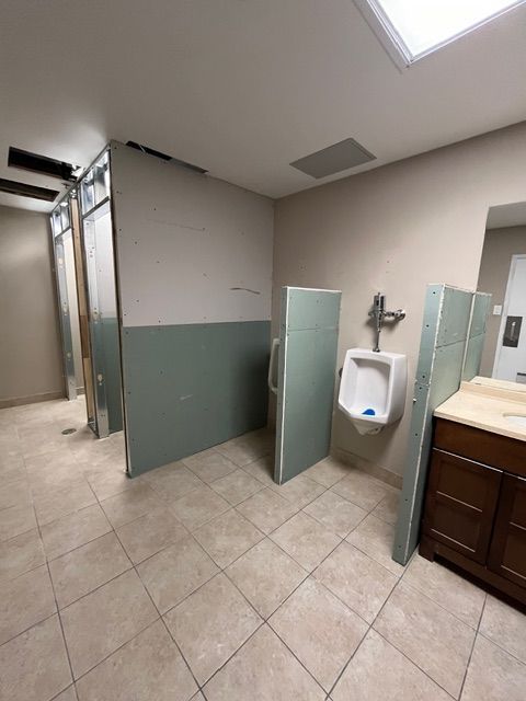 Men's restroom under renovation; exposed walls, partially constructed stalls, urinal, and sink area. Light beige walls, green partition walls.