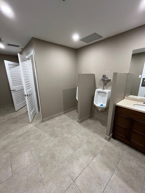 Public restroom with two urinals, two stalls, and a vanity. Light brown walls and tile floors.