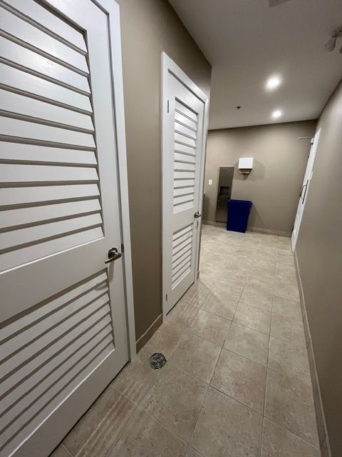 Hallway with beige walls, tiled floor, and two white louvered doors; a mirror, and a blue trash bin.