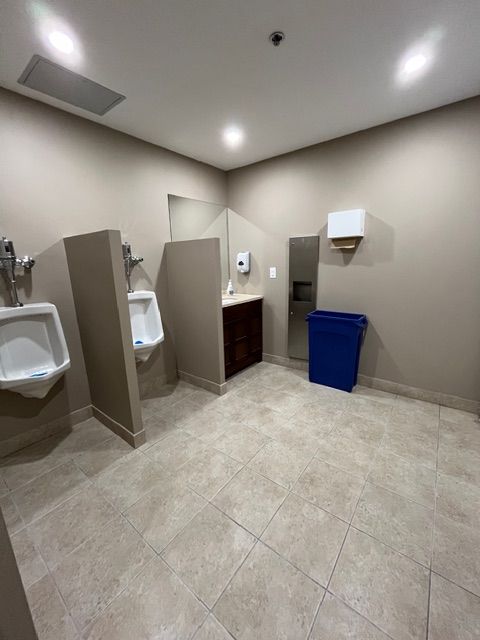 Public restroom with two urinals, a sink, and a trash can. Tile floor, beige walls.
