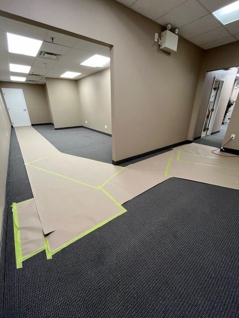 An empty office interior with dark carpet, tan walls, and a protective paper walkway outlined with green tape.