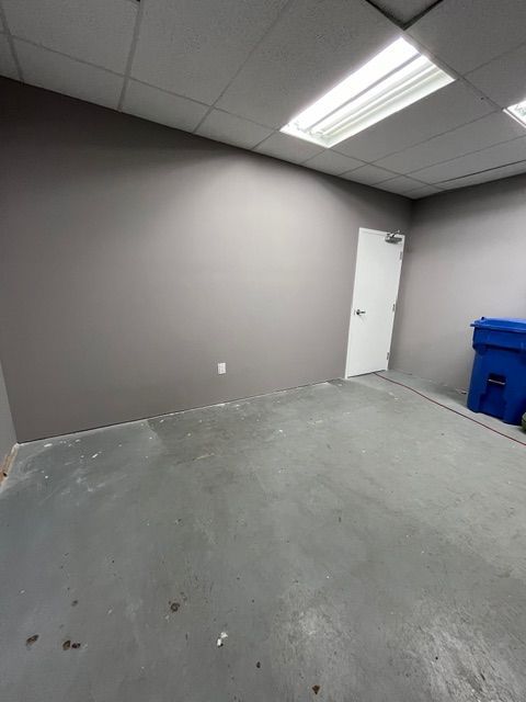 Empty room with gray walls and floor, white door, fluorescent lights, and a blue bin.