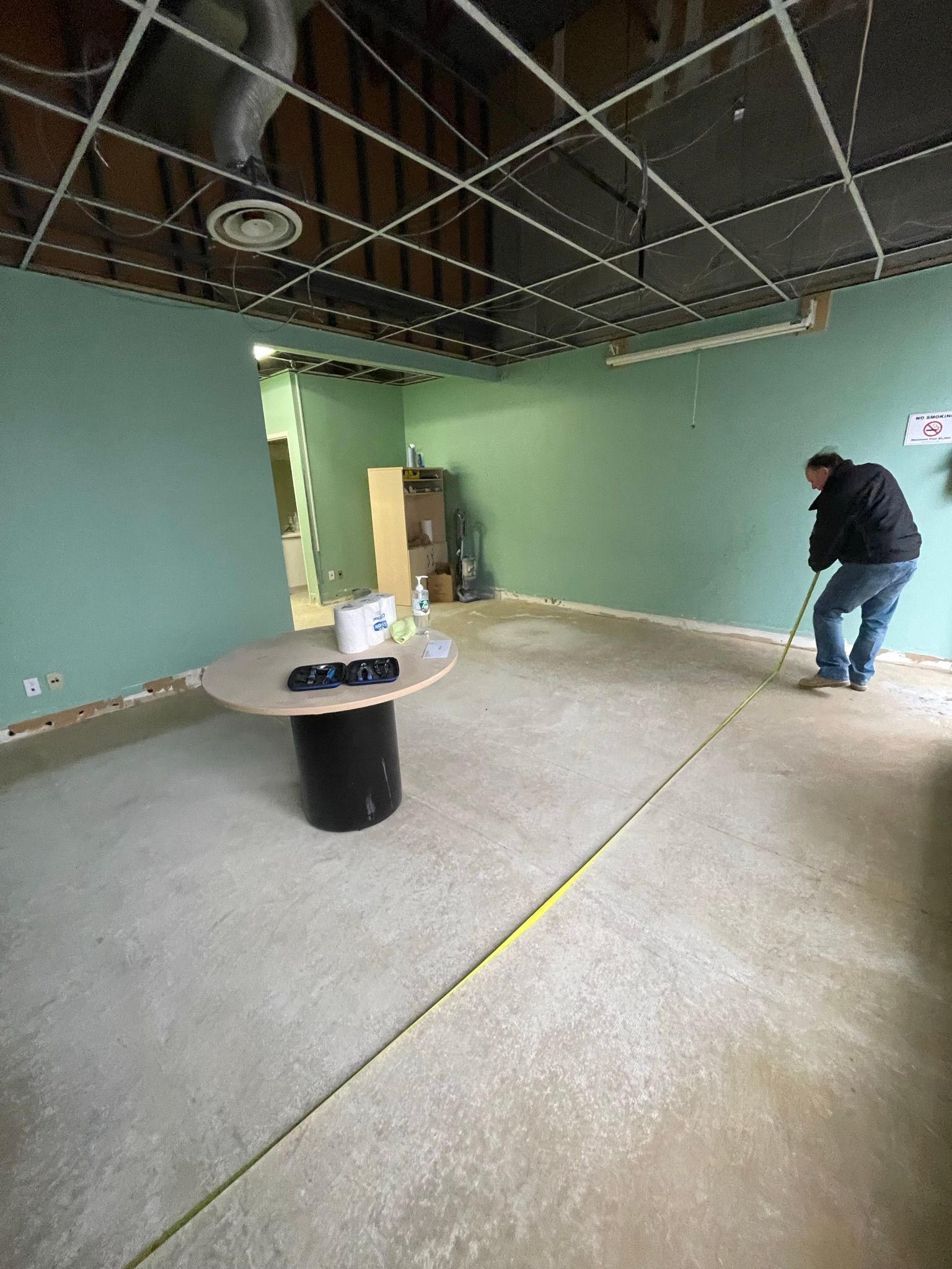 Person measuring a room with a tape measure; the walls are green, unfinished ceiling, a table is present.