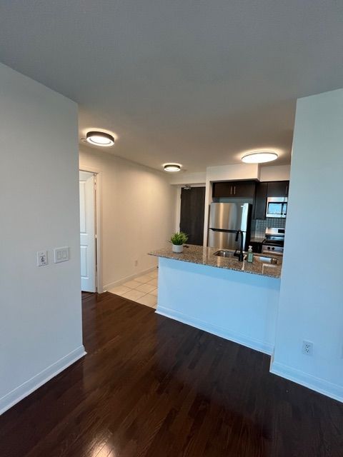 Interior view of a modern apartment with dark wood floors, white walls, and a kitchen with stainless steel appliances.