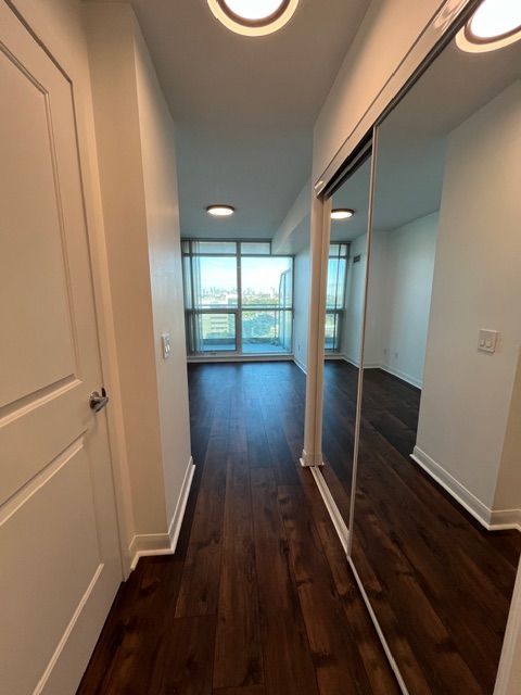 Hallway with dark wood flooring, white walls, and a mirrored closet, leading to a room with large windows.