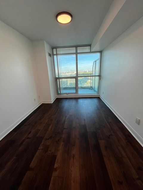 Empty room with dark wood flooring, white walls, and a large window with city view.