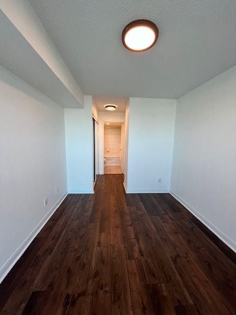 Empty room with dark wood floor, white walls and ceiling, two lights, hallway with bathroom visible.