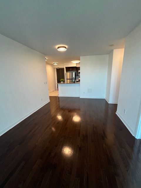 Empty apartment with dark hardwood floors, white walls, and a glimpse of a kitchen.