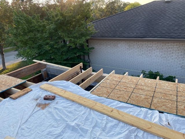 Roof construction with exposed wooden beams, plywood, and a tarp. Brick and sunlight visible.