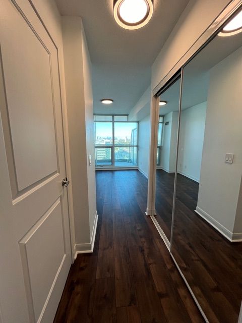 Narrow hallway with dark wood flooring, mirrored closet doors, and a window at the end.