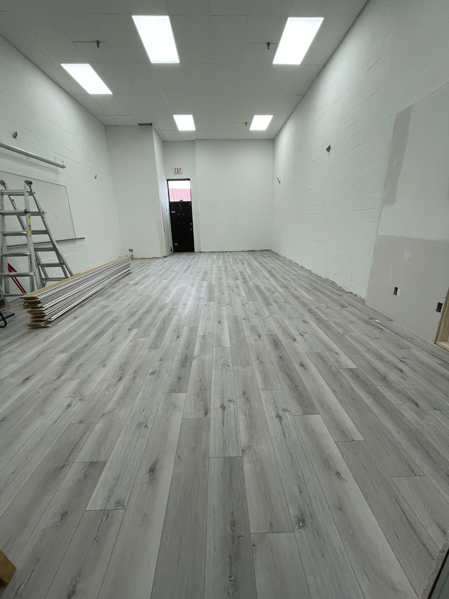 Empty commercial space with light gray wood-look flooring, white walls, and bright rectangular ceiling lights.