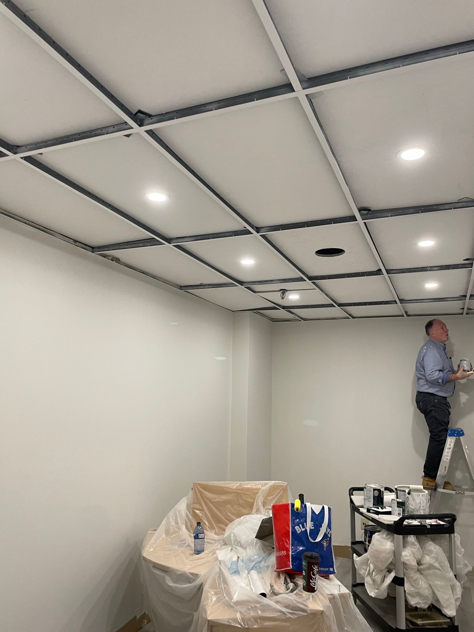 Person on ladder painting a white wall in a room with a grid ceiling and recessed lights. Paint supplies on covered table.
