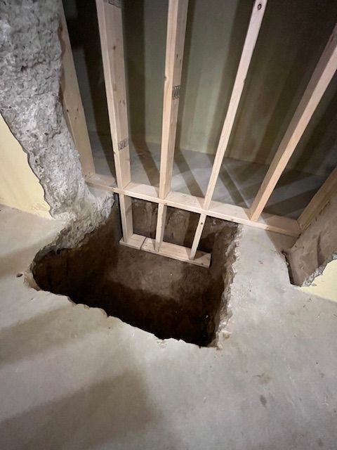 Hole in a concrete floor, framed with wood, revealing an excavated space below, likely for access or utilities.
