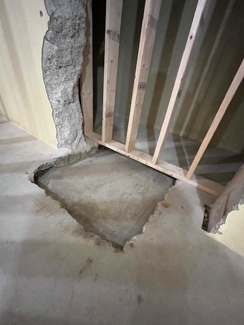 Hole cut in concrete floor and wall, revealing unfinished wooden framework; interior shot.