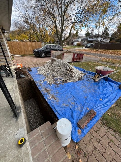 Construction site: trench dug next to house, covered with blue tarp, gravel pile, wheelbarrow, and bucket.
