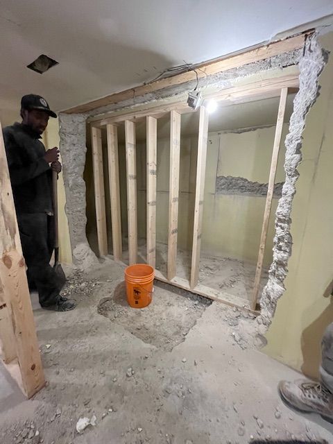 Man building a wooden wall frame in a room under construction. Orange bucket sits on the floor.