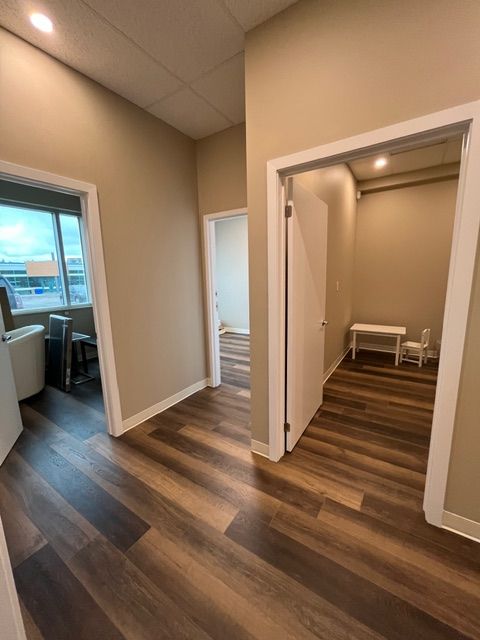 Hallway with three open doors, brown and tan walls, wood-look flooring. One door shows a desk, another a small table and chairs.