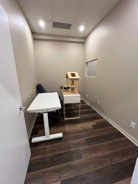 Small room with a desk, chair, and decorative wooden house. Dark wood floor, beige walls, and white door.