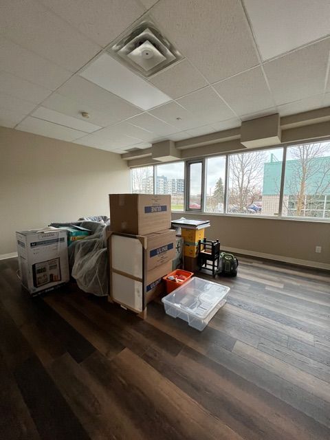 Empty room with moving boxes near a window, brown floor, tan walls, and a white ceiling with vents.