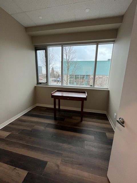Empty room with dark wood flooring, small wooden desk, and large window.