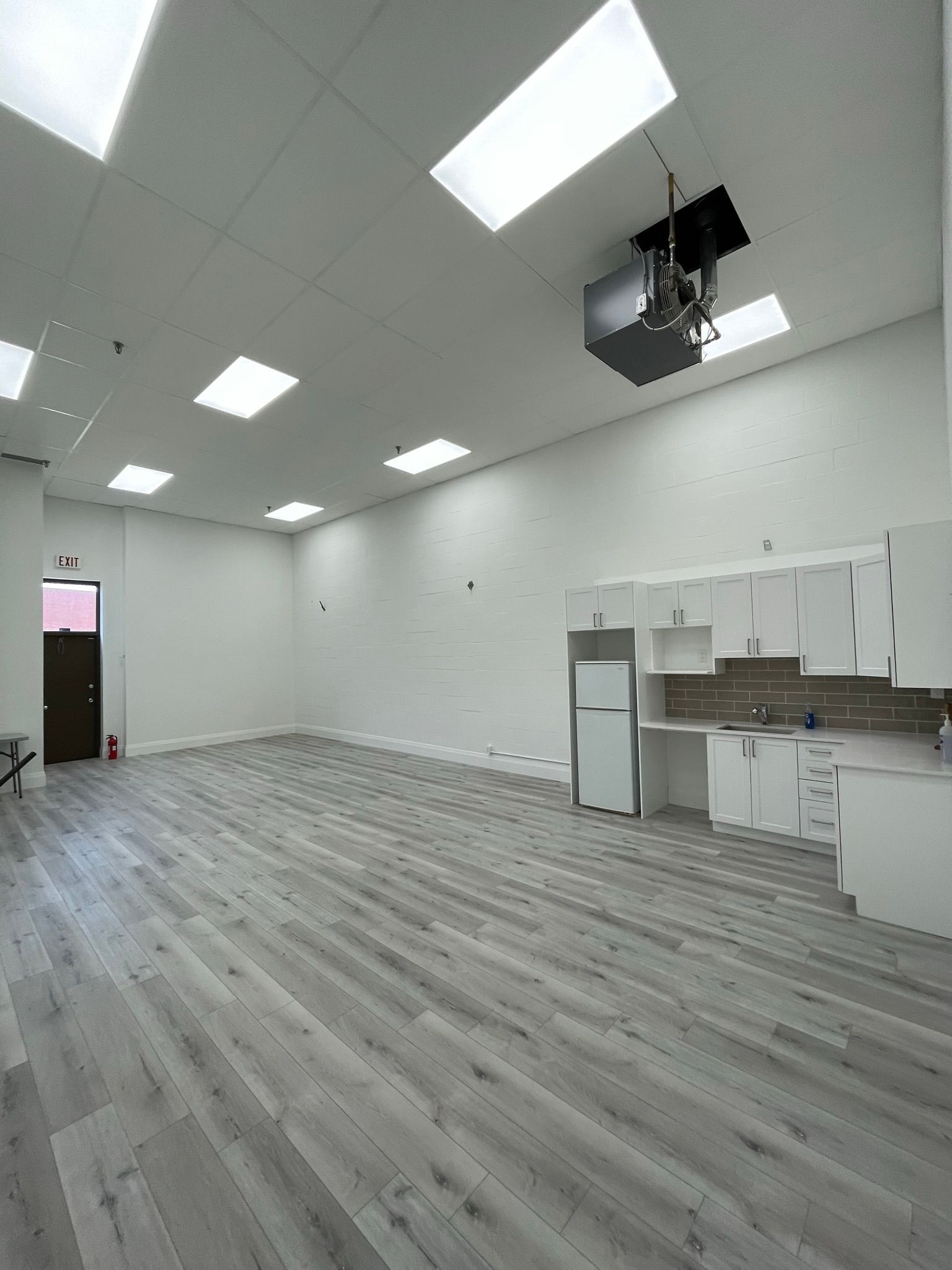 Empty commercial space with white walls, gray wood-look floor, and a kitchenette.