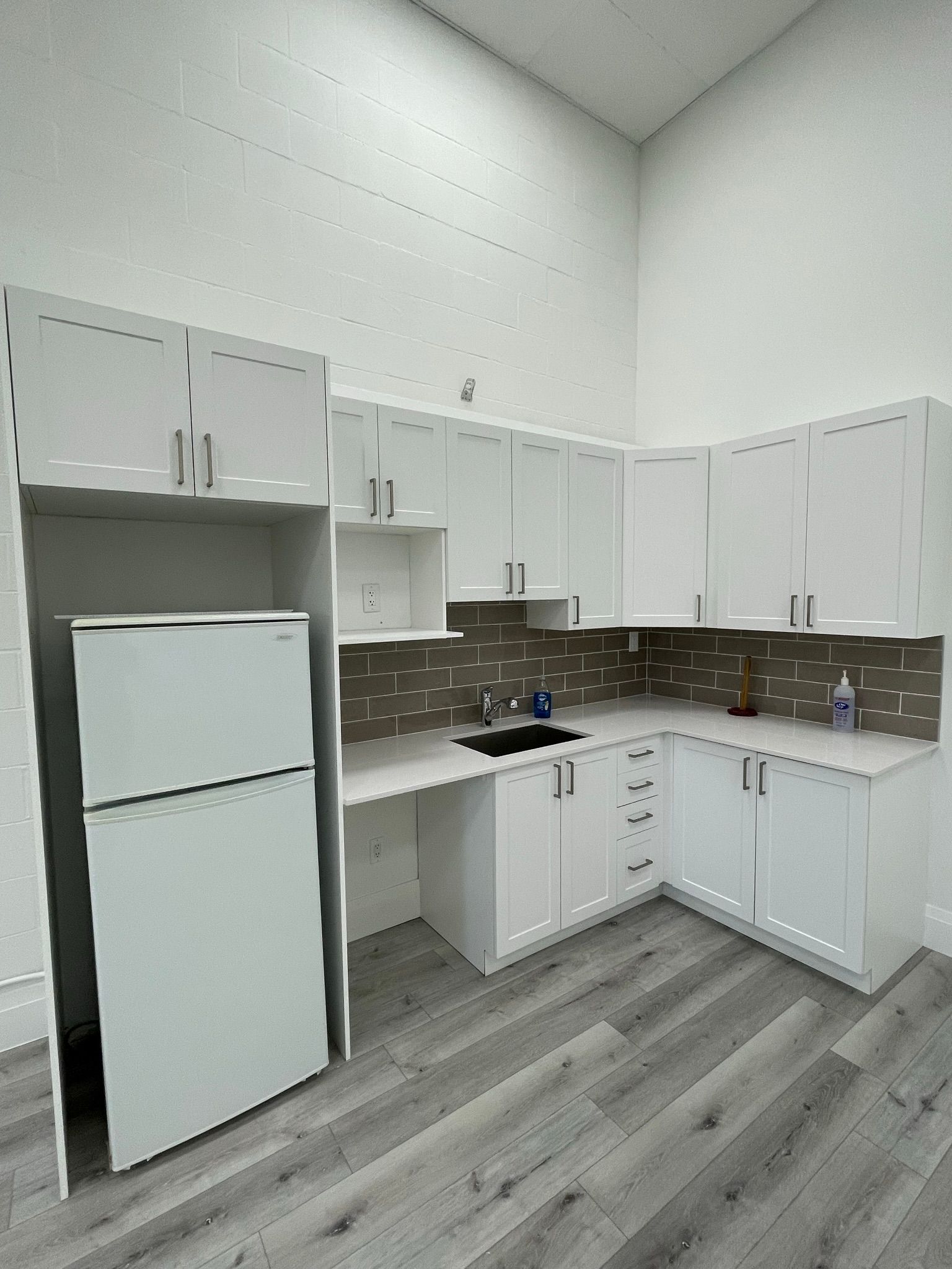 White kitchen with cabinets, refrigerator, and countertop. Dark backsplash, grey flooring, and high ceilings.